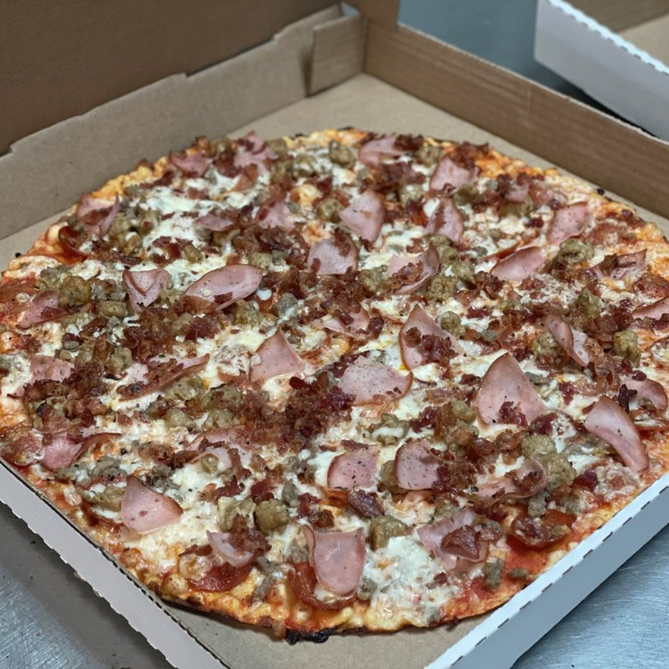 A Lion's Den Pizzeria meat lover's pizza, topped with ham, sausage, and bacon, in a cardboard box.