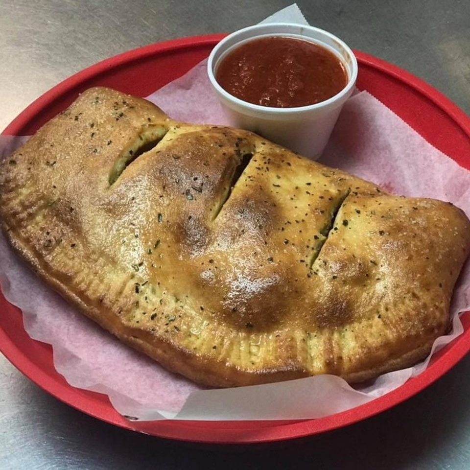 Golden-brown calzone with herbs and cuts, served with a side of marinara sauce in a red basket.