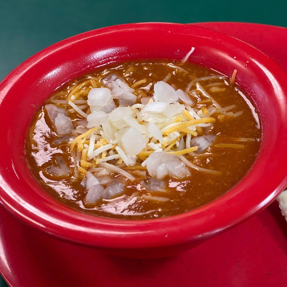A red bowl of chili topped with shredded cheese and diced onions.