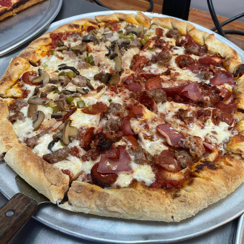 A half-and-half Lion's Den Pizzeria pizza with meat and vegetable toppings on a metal tray.