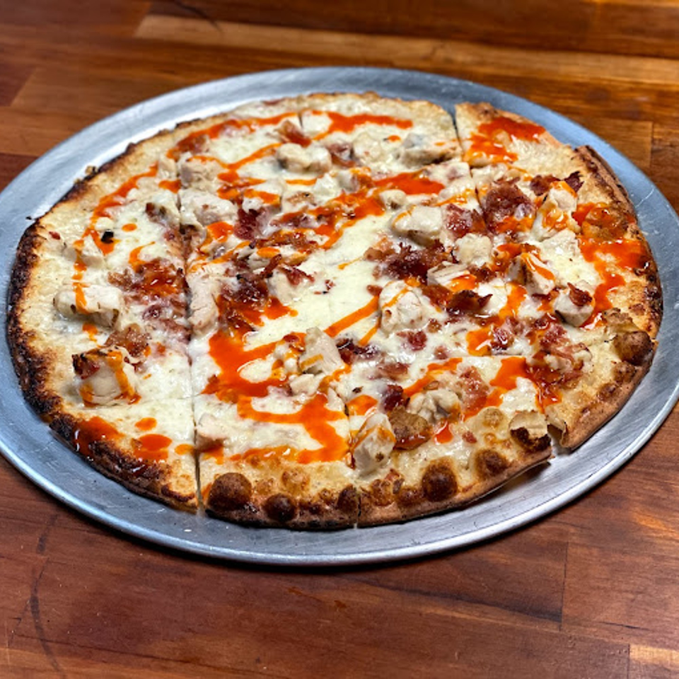 A Buffalo chicken pizza from Lion's Den Pizzeria, with white sauce, chicken, bacon, and hot sauce drizzle.