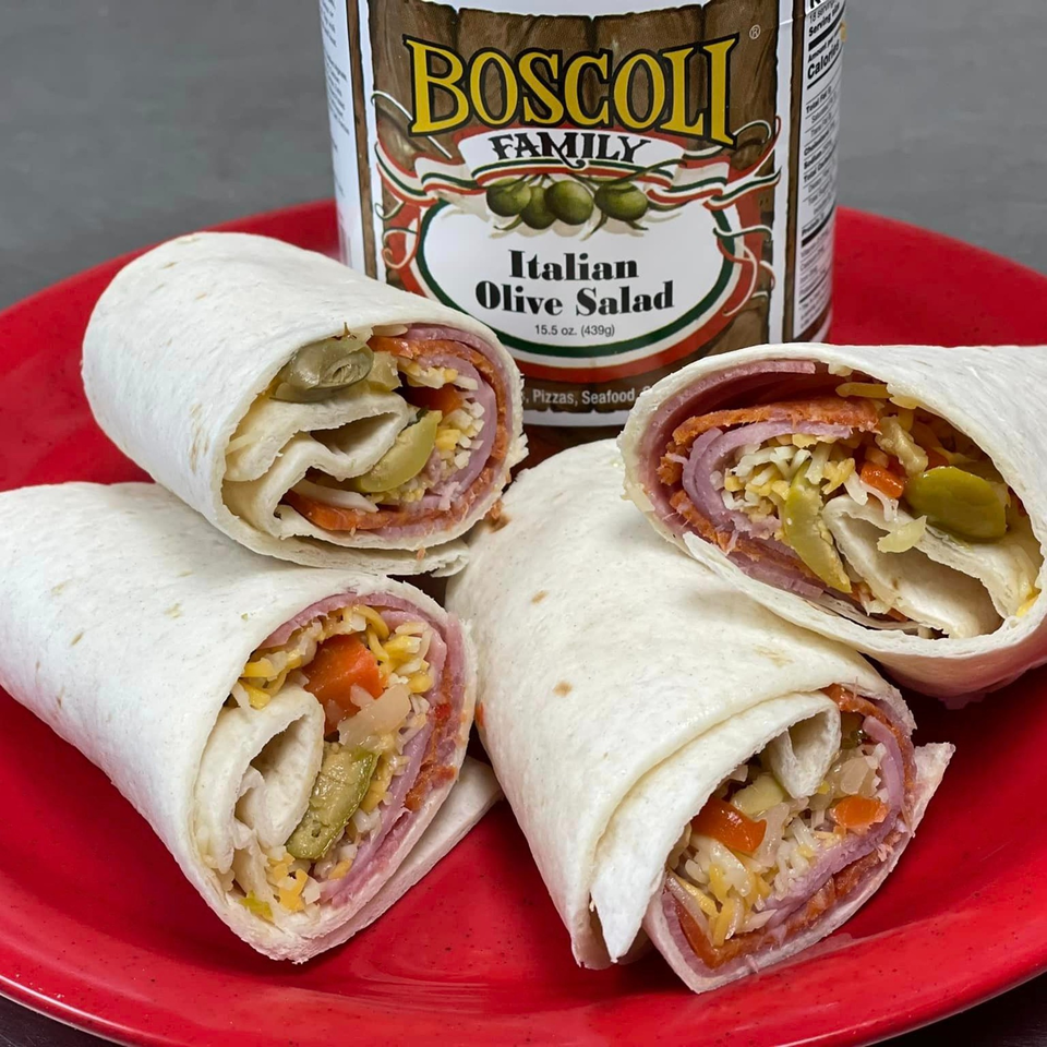 Four Italian wraps filled with meats, cheese, and olive salad, on a red plate with a jar of Boscoli Italian Olive Salad.