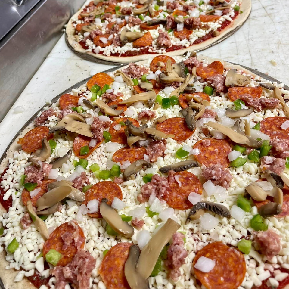 Two uncooked pizzas from Lion's Den Pizzeria, topped with pepperoni, sausage, mushrooms, onions, and green peppers.